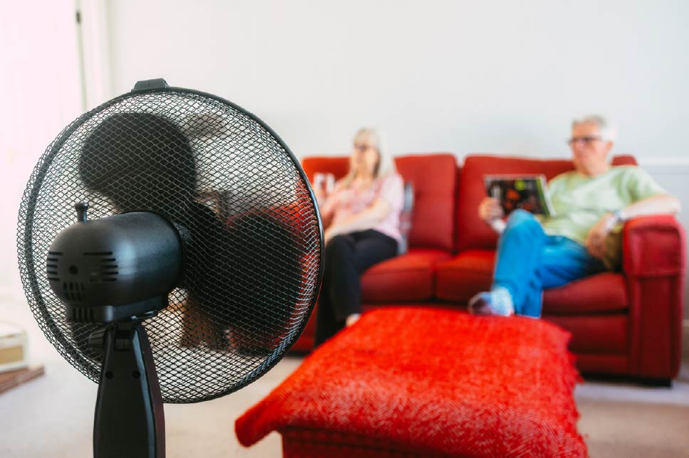 A couple sitting on a red sofa with a large fan keeping them cool.
